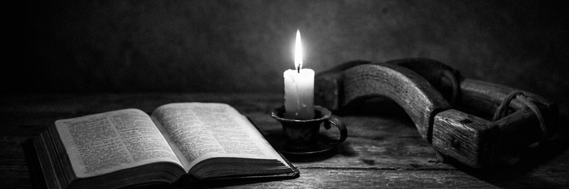 Black-and-white still life of an open Bible on a wooden table beside a lit candle and a wooden cross, symbolizing quiet faith and prayer.